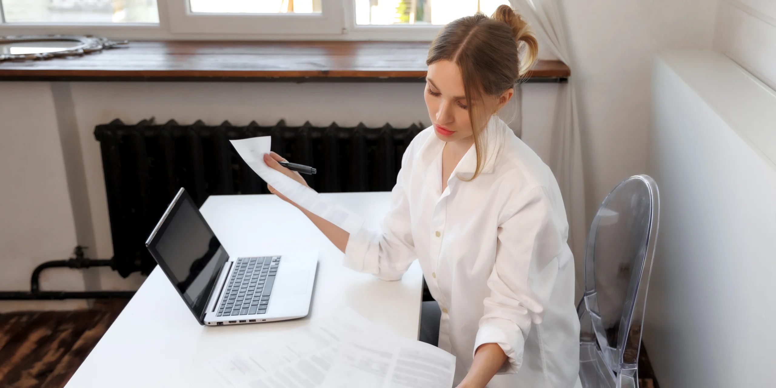 A woman in a white shirt reads through printed pages at a white desk by a window, her laptop open beside her