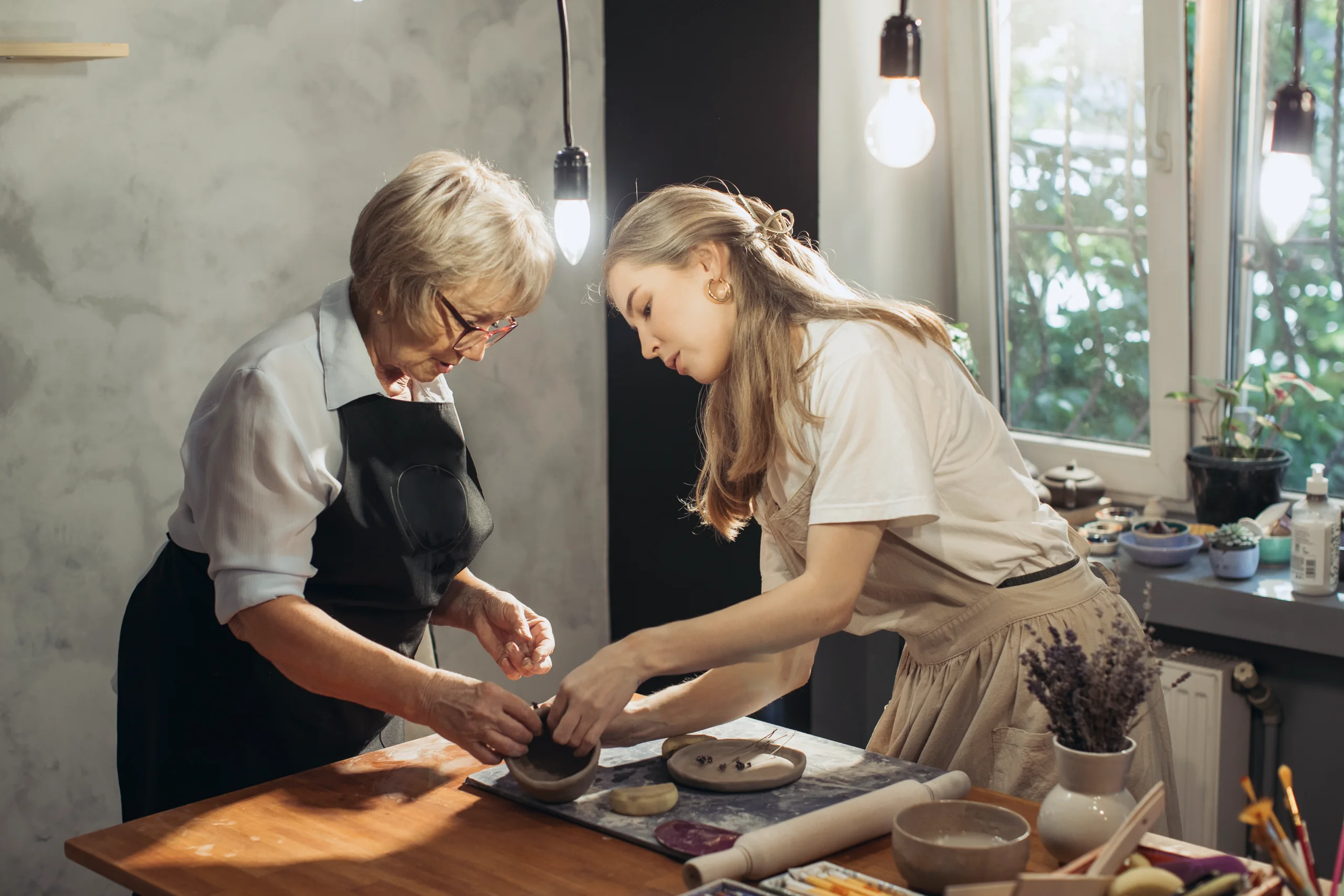 An older potter and a younger woman shape a small clay bowl together, their hands meeting over it at a wooden worktable by a sunlit window